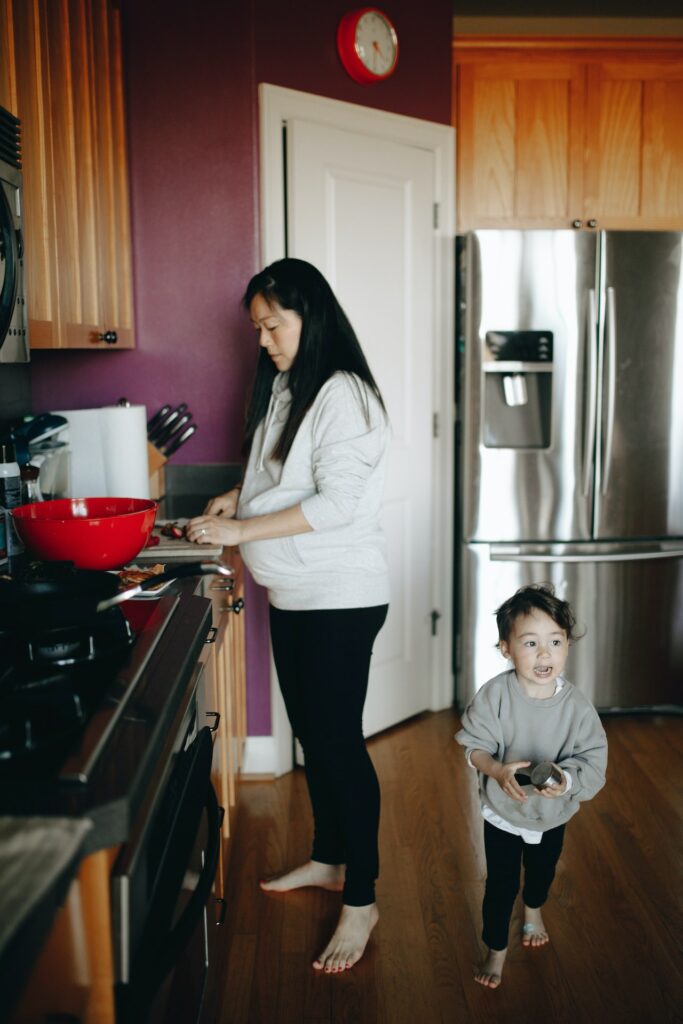 身穿白运动衫和黑腿的孕妇在孩子附近割菜,厨房内穿灰运动衫和黑腿